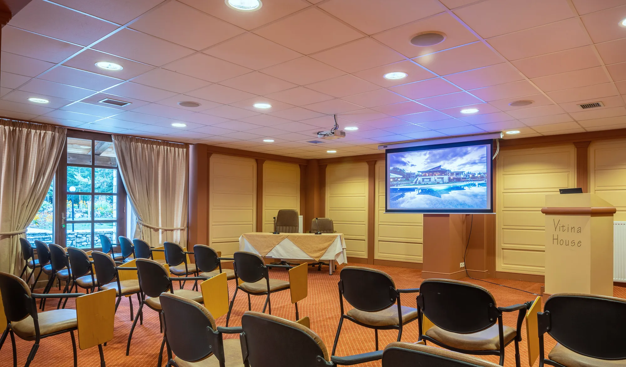 The conference room set with rows of chairs facing a projection screen and a lectern, daylight coming through windows on the left
