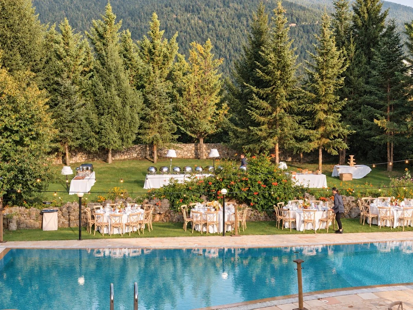 Round tables set on the lawn and buffet tables under the firs, beside the pool, in the afternoon sun