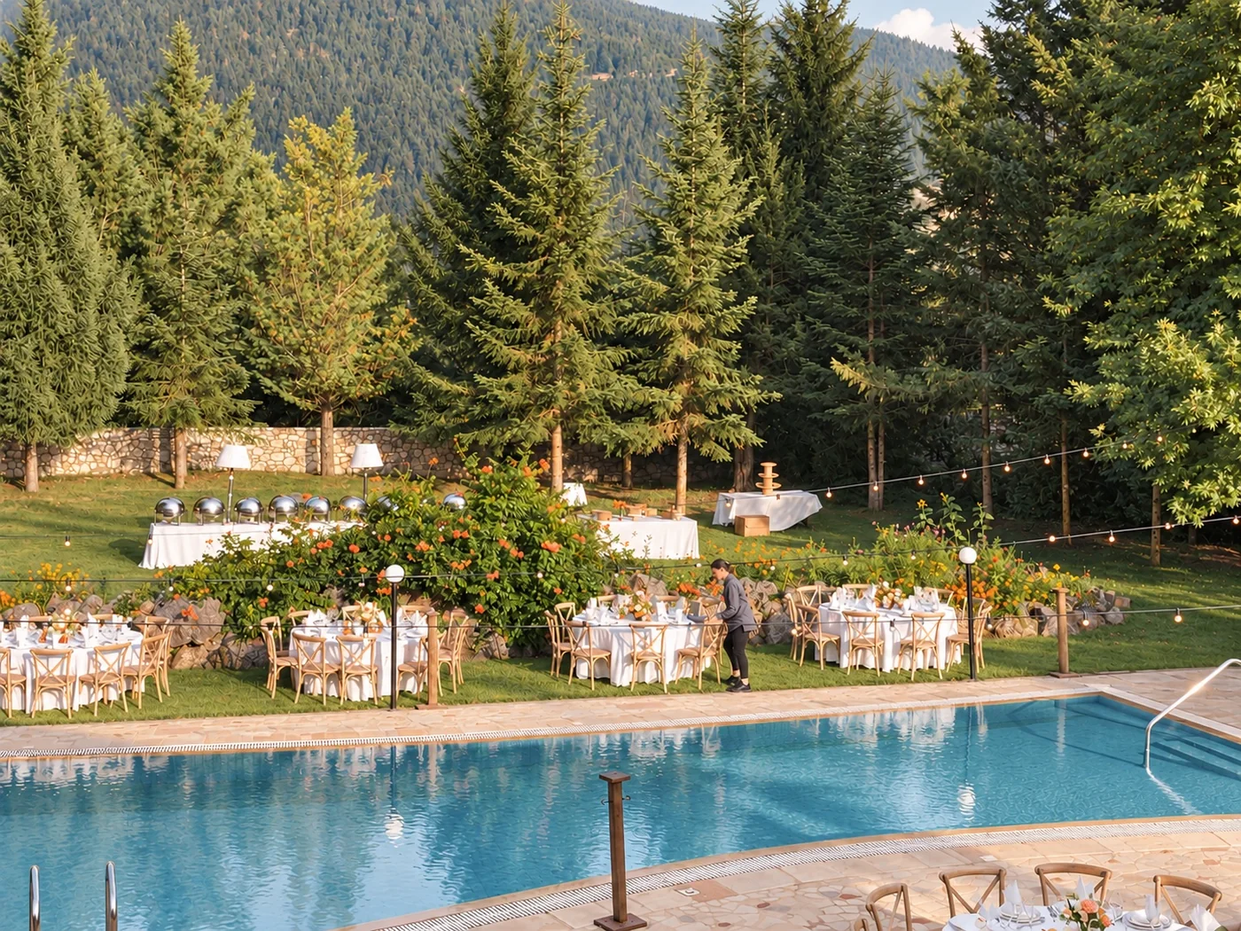 Round tables with chairs on the grass behind the pool, with string lights and firs above