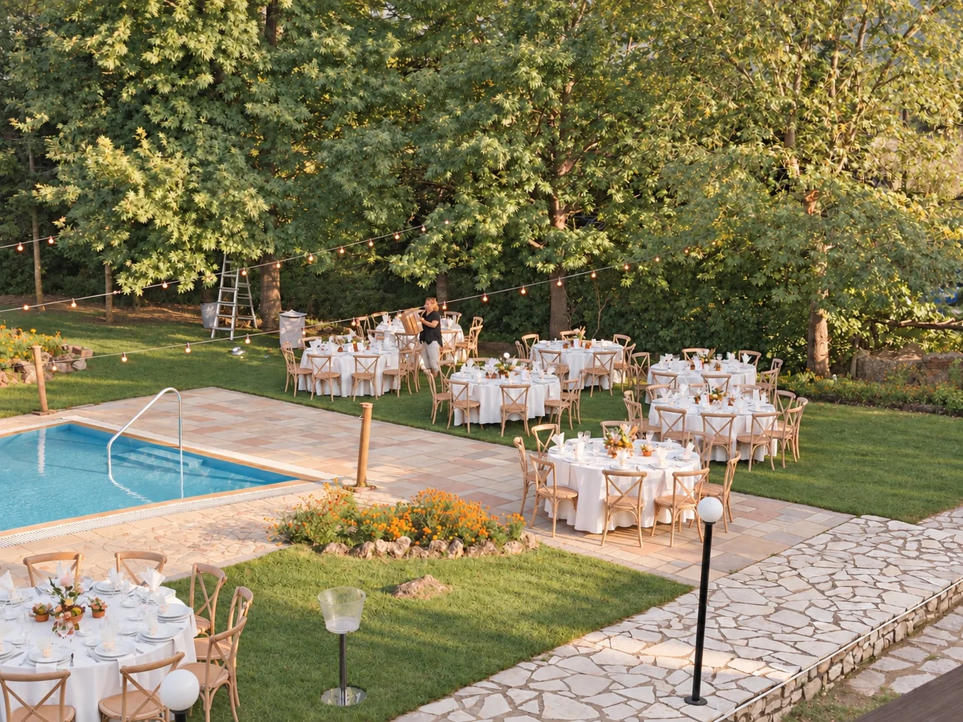 Round tables laid with plates and flowers on the lawn under tall trees, with string lights
