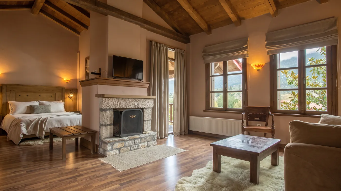 Superior room with a stone fireplace, timber beams and tall windows facing the forest