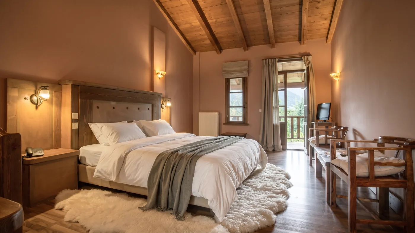 Standard room: a bed with linen cover and a sheepskin rug, timber ceiling, and a balcony door onto the mountains