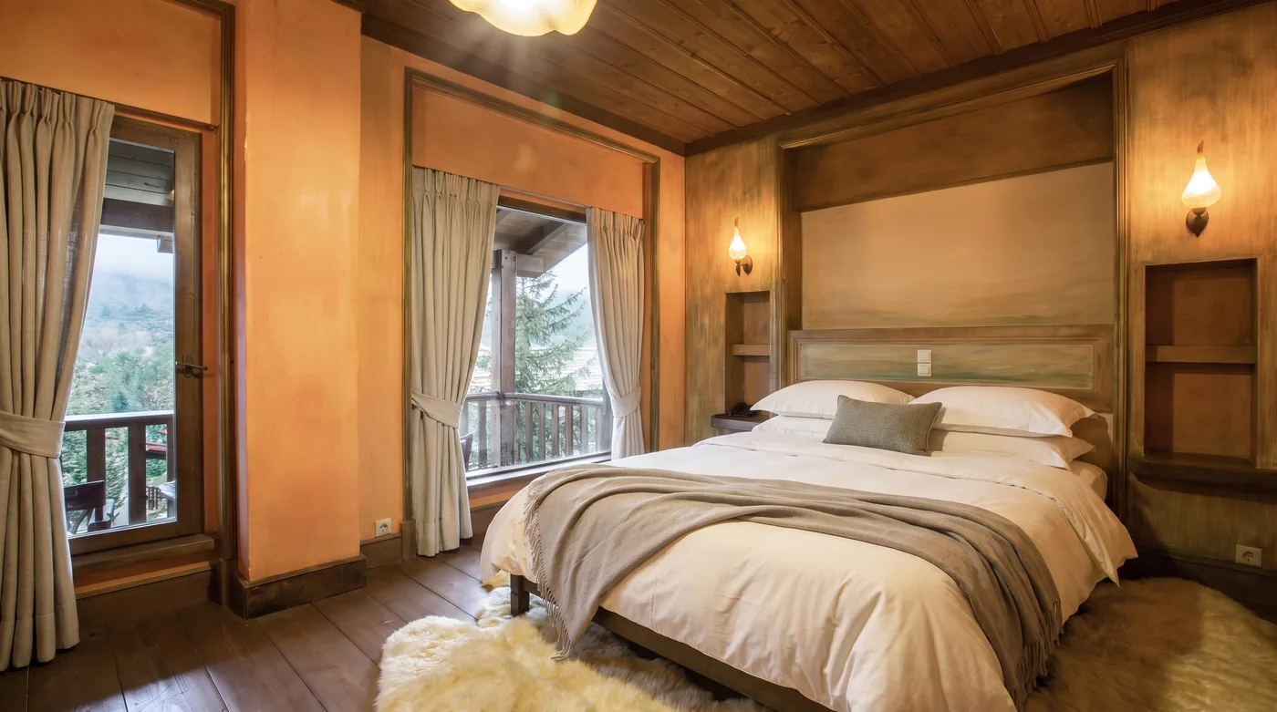 Suite bedroom with a king-size bed in white linen and a taupe throw, a carved timber headboard recess, wall lamps and a balcony door onto the mountain view