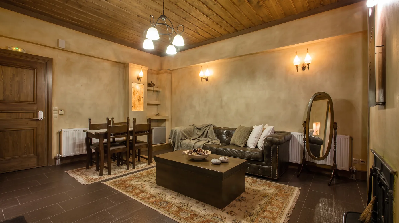 Suite living room with a leather sofa, a wooden chest used as a coffee table, a dining table with chairs and a patterned rug under a timber ceiling