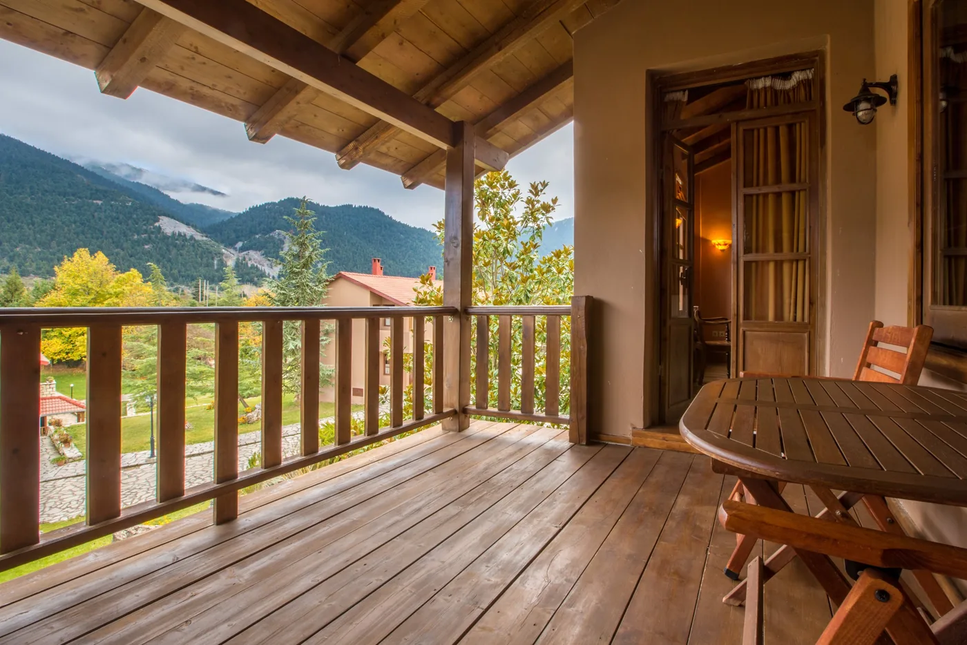 Covered timber balcony with a table and chairs, an open door to the room, and the garden and forested mountains beyond