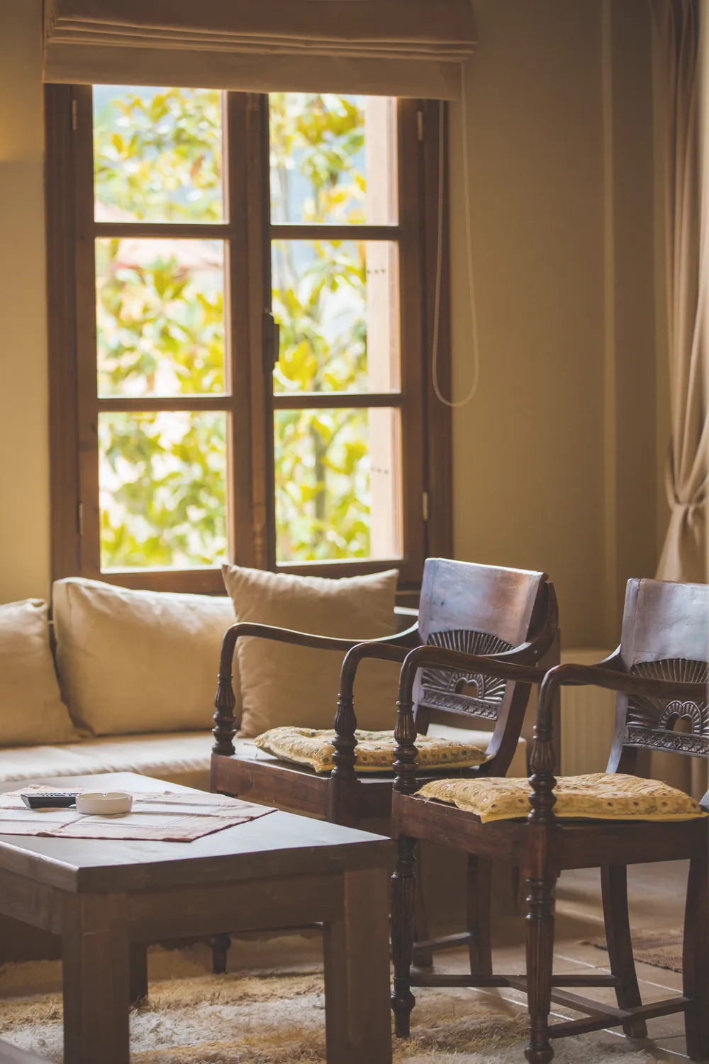 Two wooden armchairs and a small table beside a tall window with green leaves outside
